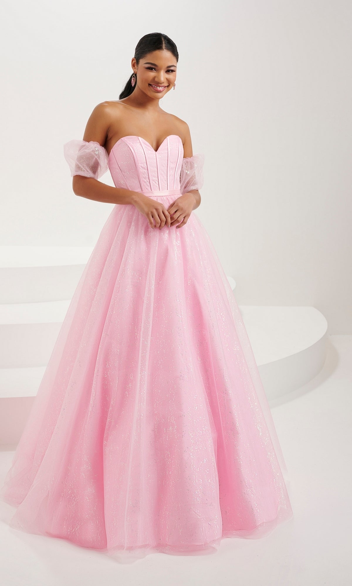 A woman stands in a white studio wearing the Tiffany Strapless Glitter Pastel Prom Dress 16083, featuring a corset bodice and sheer off-the-shoulder sleeves.